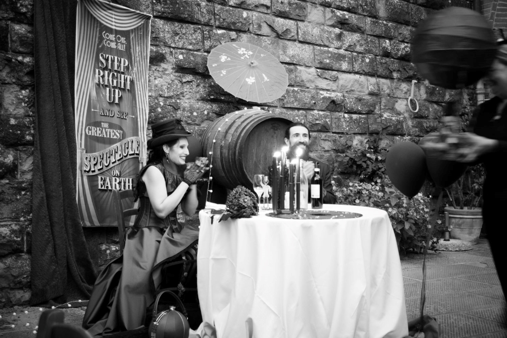 A black and white photograph of two laughing spouses at a table, with a victorian circus banner and a wine barrel behind them