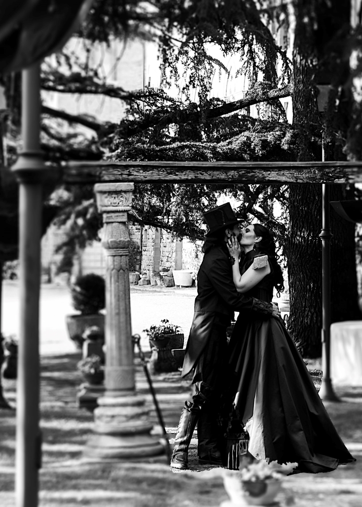 A black and white photo of two spouses dressed in a gothic fantasy style, kissing under an archway