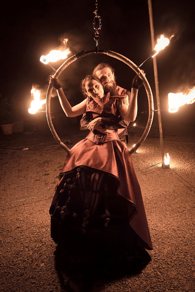 A full figure photo of spouses in gothic fantasy clothing, posing inside a fire circle and smiling