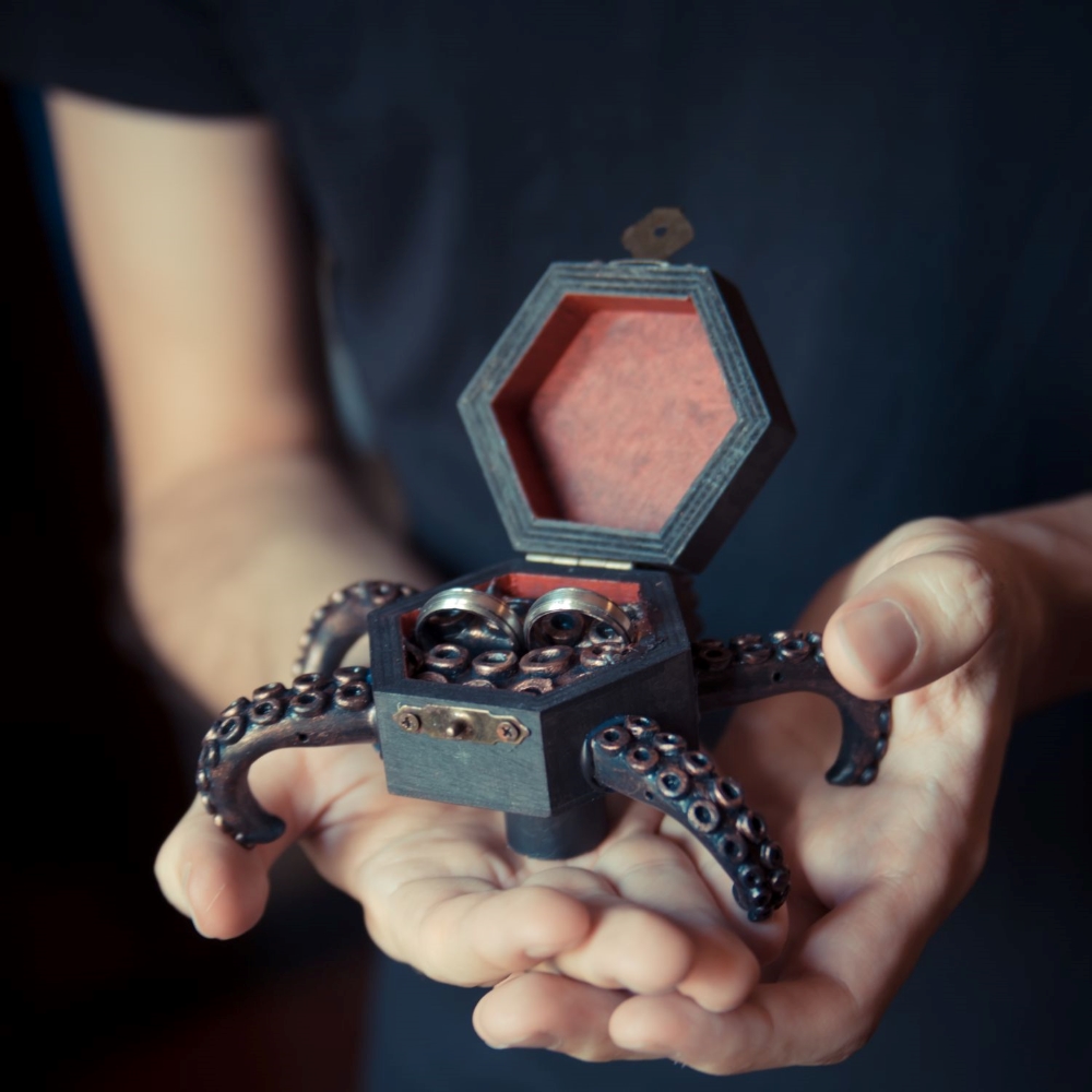 Hands holding up a red and black wedding rings box with octopus arms, in the style of H.P. Lovecraft