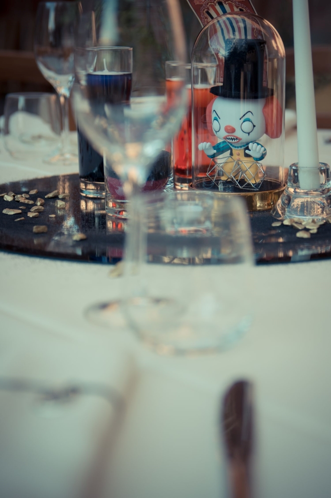 A steampunk centerpiece of an evil clown similar to IT, under a glass dome, surrounded by candles floating in water glasses