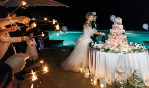 The spouses in their perfect venue, cutting the cake in front of the swimming pool with people all around with magic sparkles