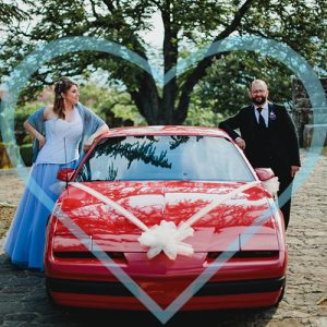 A couple of spouses looking happy as they lean on their red sports car, with a blue heart on top